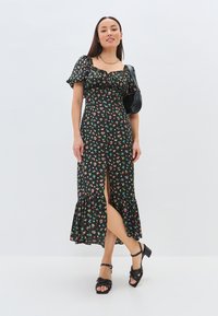 Robe noire à fleurs avec de courtes manches bouffantes, ourlet froncé et fente sur le devant. Présente des fleurs multicolores et un corsage ajusté. Sandales à talons noires à lanières.