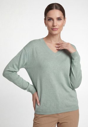 Vrouw met lichtgroene V-hals trui en beige broek, poserend met één hand in de taille en de andere bij haar sleutelbeen, neutrale achtergrond.