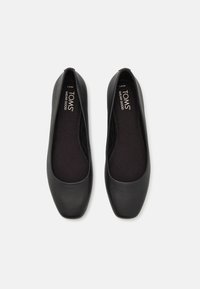 Zapatos planos negros con un acabado de cuero suave, punta redondeada y plantilla acolchada. Logo de TOMS impreso en las plantillas. La suela tiene una textura mínima.