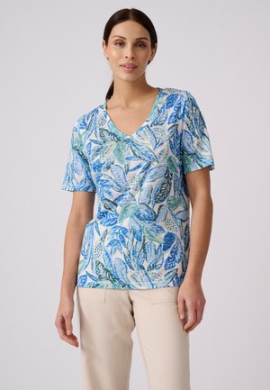 Femme aux cheveux foncés portant un haut bleu et vert à motif de feuilles, manches courtes, col en V, et un pantalon beige clair, debout devant un fond uni.