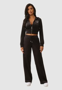 Ensemble de survêtement en velours noir comprenant un hoodie zippé court et un pantalon à jambes larges avec taille à cordon. Tissu texturé, détail logo subtil sur la poitrine.
