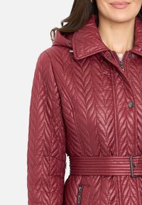 Chaqueta burdeos acolchada con un alto cuello, cintura ceñida con cinturón y cremalleras laterales. La costura texturizada crea un patrón de hojas en la superficie.