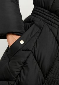 Giubbotto puffo nero con motivo a rombi diagonali, dotato di tasca laterale chiusa da un bottone a pressione dorato e vita elastica arricciata.