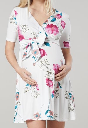 Zwangere vrouw in een witte jurk met roze en blauwe bloemen, handen rusten zachtjes op haar buik, staand tegen een grijze achtergrond.