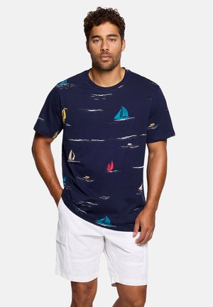 Hombre con camiseta azul marino con estampados coloridos de veleros y pantalones cortos cargo blancos, de pie con las manos en los bolsillos.