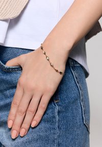 Bransoletka w złotym odcieniu z wydłużonymi, teksturowanymi ogniwami w kształcie liści, noszona na dłoni spoczywającej na kieszeni dżinsów. Widoczna gładka skóra i jasny lakier do paznokci.