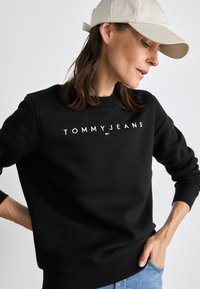 Felpa nera in misto cotone con collo rotondo, contraddistinta dal logo "TOMMY JEANS" in bianco, abbinata a un cappellino beige chiaro.