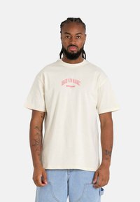 Crème-kleurig t-shirt met korte mouwen en een ronde halslijn, met een rood bedrukte logo. Heeft een losse pasvorm en een gestructureerde stof.