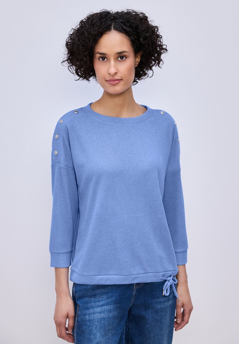 Femme aux cheveux bouclés portant un pull bleu clair avec des détails de boutons sur les épaules et un jean bleu, debout devant un fond uni.