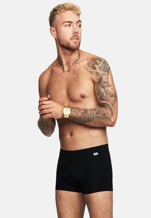 Black boxer briefs made of stretchy fabric, featuring a smooth waistband and minimal branding. Model has visible tattoos and a watch.