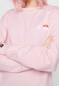 Ljusrosa sweatshirt med broderad "elless"-logotyp och färgstark detalj. Långa ärmar med subtil textur och märkt mudd.