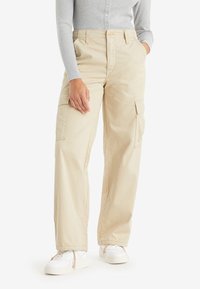 Osoba v béžových širokých kalhotách s cargo kapsami, šedém pleteném dlouhém topu s dlouhým rukávem a bílých teniskách, stojící s jednou rukou v kapse.