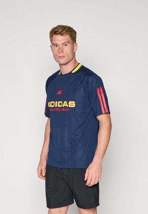 Camiseta deportiva azul marino con cuello amarillo y detalles rojos. Presenta un gran logo "ADIDAS" en amarillo y pantalones cortos negros. Textura suave, diseño minimalista.