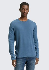 Langärmeliger blauer Baumwollpullover mit strukturiertem Rippstrick, runder Ausschnitt, entspannte Passform, getragen mit verwaschenen Jeans.