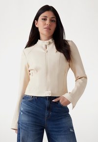 Béžová zkrácená bunda s stojáčkem, předním zipem a zapínáním na dva knoflíky. Dlouhé rukávy s přiléhavými manžetami; hladká, koženková textura.