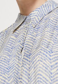 Camicia a maniche lunghe con una base gialla chiara e un motivo a zigzag blu. Presenta un colletto classico e una texture di tessuto morbido.