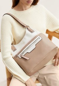 Bolso de hombro en tonos marrón y blanco con forma redondeada, material de cuero, acentos en contraste, cierre superior de cremalleras y un bolsillo frontal decorativo.
