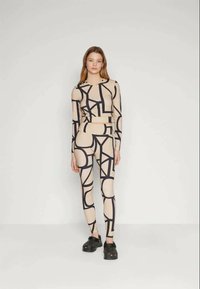 Beige crop top és leggings, amelyek merész fekete geometrikus mintákkal díszítettek, fekete klumpákkal párosítva. Puha, rugalmas anyag, amely testhezálló fazonú.