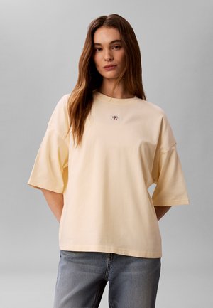 Junge Frau mit langen braunen Haaren, die ein übergroßes beiges T-Shirt mit kleinem Logo und blaue Jeans vor einem schlichten grauen Hintergrund trägt.