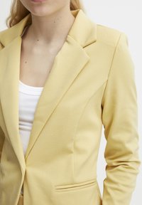Blazer jaune à coupe ajustée, avec des revers nets et une texture lisse. Porté sur un haut blanc, il comporte une poche frontale unique.