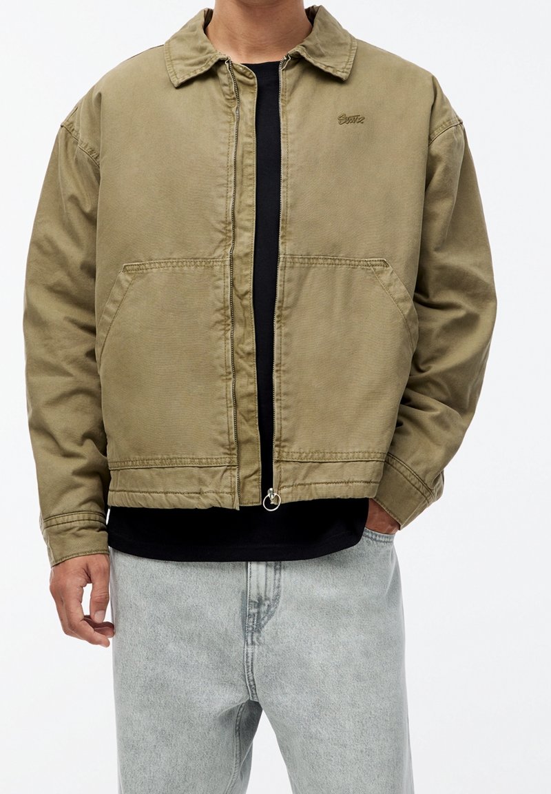 Veste bomber khaki avec fermeture éclair, col rabattu et deux poches avant. Portée sur un haut noir et associée à un jean gris clair.