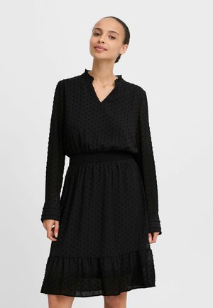 Black dress with long sleeves, V-neck, textured polka-dot fabric, smocked waist, and flared ruffled hem. Semi-sheer material.