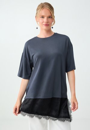 Młoda kobieta ubrana w ciemną luźną bluzkę z krótkim rękawem, z czarną koronką na dole, nałożoną na białe spodnie, stojąca na jednolitym jasnym tle.