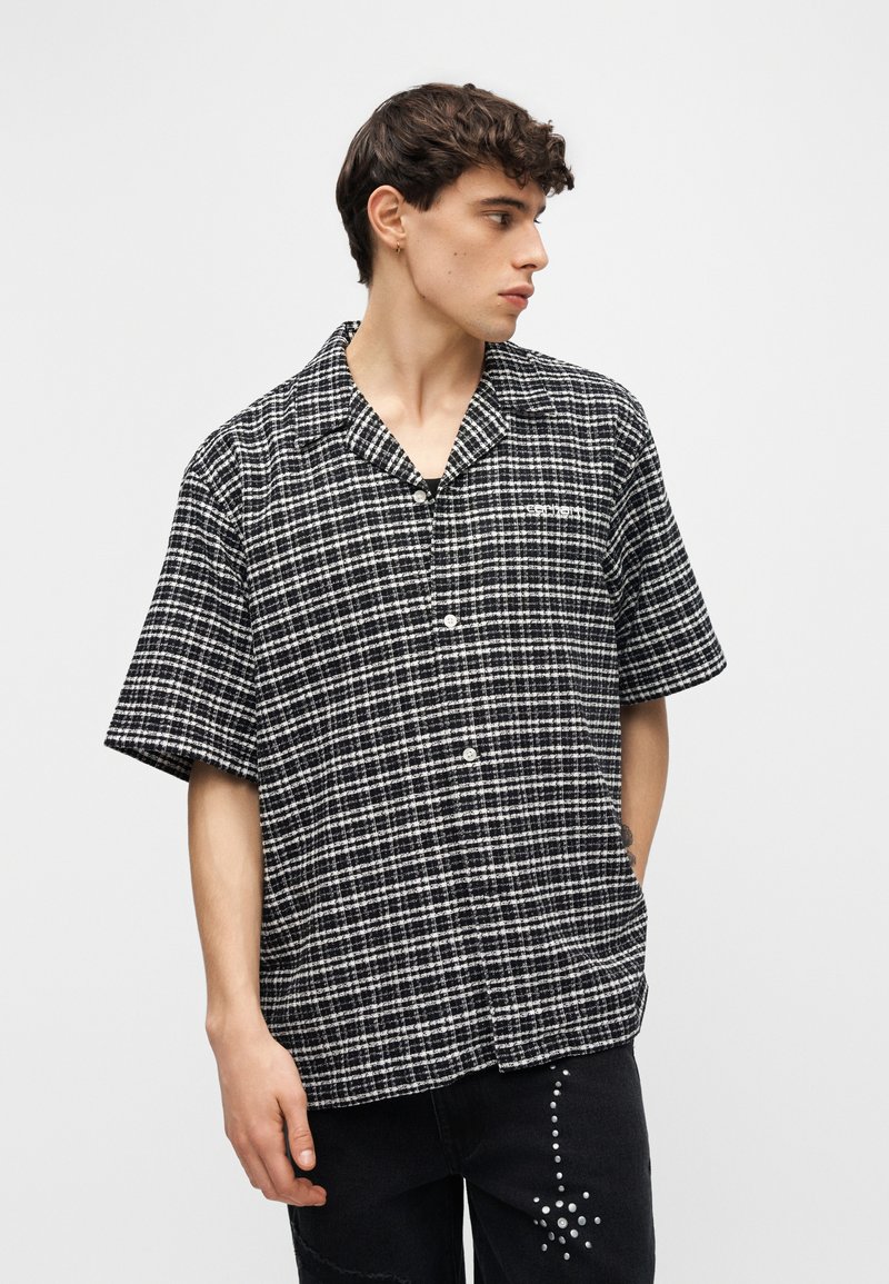 Giovane che guarda a destra, indossa una camicia a maniche corte con bottoni a quadri bianchi e neri e pantaloni neri con design a stelle bianche a pois.