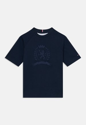 Camiseta azul marino con cuello redondo y mangas raglán cortas. Presenta un diseño de emblema bordado y un pequeño logotipo en la manga.
