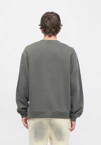 Felpa grigia con vestibilità comoda, polsini a coste e scollo rotondo. Materiale in misto cotone, vista posteriore senza dettagli, indossata con pantaloni di colore chiaro.