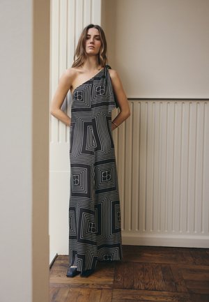 Femme debout à l'intérieur, portant une combinaison noire et blanche une épaule avec des motifs géométriques carrés et des chaussures noires.