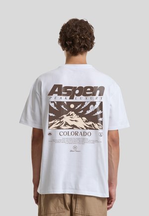 Personne avec des cheveux bouclés porte un t-shirt blanc avec un motif marron d'une montagne, des nuages et le texte "Aspen Peak Luxury Colorado" dans le dos.