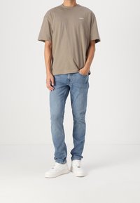 Jasnobrązowy bawełniany t-shirt z okrągłym dekoltem i krótkimi rękawami, w połączeniu ze średnio-niebieskimi dżinsami i białymi trampkami.