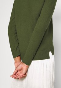 Olivgrön stickad tröja med ribbade ärmslut, sidofickor och en mjuk textur, kombinerad med en plisserad vit kjol. Visas från baksidan.