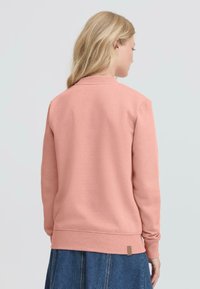 Lys rosa hettegenser med ribbestrikket krave, lange ermer og teksturert stoff. Har en liten brun merkelapp ved kanten.