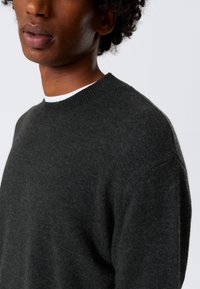 Grå strikkegenser med rund hals. Stoffet ser mykt og teksturert ut, med en tettsittende design. En hvit skjorte er lagdelt under.