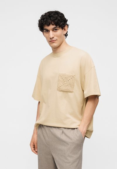 Νεαρός άνδρας με σγουρά σκουρόχρωμα μαλλιά, φορώντας μπεζ φαρδιά μπλούζα με υφή στην τσέπη και ανοιχτό γκρι παντελόνι, στέκεται με το ένα χέρι στην τσέπη.