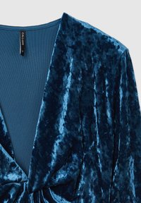 Top avvolgente in velluto di un blu profondo con una texture liscia. Presenta un design a V bassa e un dettaglio annodato plissettato sul davanti, creando un effetto drappeggiato.