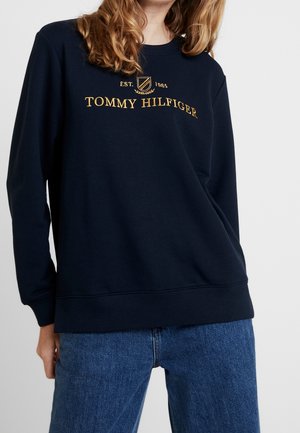 Person trägt einen dunkelblauen Pullover mit gelbem "Tommy Hilfiger EST. 1985" Logo und blaue Jeans, steht vor einem einfarbigen hellen Hintergrund.
