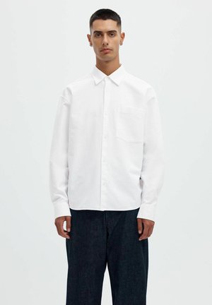 Jeune homme debout, portant une chemise blanche boutonnée à manches longues et un pantalon ample foncé sur un fond clair uni.