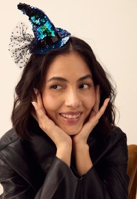 Paillettenheksenhoed in iriserend blauw en groen met zwarte rand, versierd met een zwarte tulen strik met stippen, schuin gedragen.