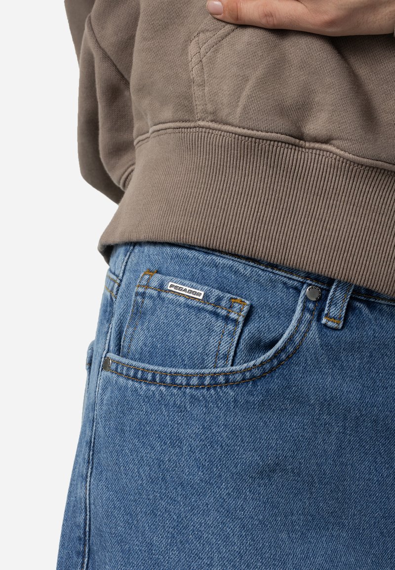Lichtblauwe spijkerbroek van denim met een vervaagde textuur, voorzien van een zilveren logopatch, zijzakken en oranje stiksels. Gecombineerd met een taupe sweatshirt.
