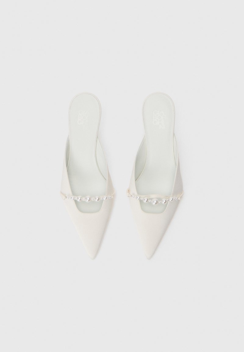 Le mule a punta bianche sono caratterizzate da una catena con perle rotonde che attraversa il dorso del piede, suola piatta e design senza schienale.