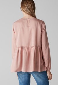 Růžová halenka s dlouhými rukávy a nařaseným peplum střihem, s uzávěrem na klíčovou dírku na zádech a hladkým textilním povrchem. Nosená s džíny.