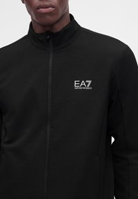 Άνδρας που φοράει μαύρο μπουφάν EA7 Emporio Armani με φερμουάρ, με υφή σχεδίου και όρθιο γιακά.