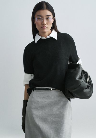 Suéter de punto negro con puños blancos doblados, llevado sobre una camisa blanca con cuello. Falda gris con cinturón, guantes negros y un bolso negro.