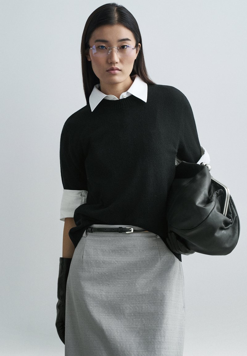 Black knit sweater with rolled white cuffs, worn over a white collared shirt. Gray skirt with a belt, black gloves, and a black bag.