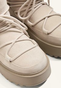 Beige Wildleder High-Top Sneakers mit dicken Sohlen, versehen mit strukturierten Akzenten und überkreuzten Schnürsenkeln. Nahaufnahme zeigt Details und Nähte.