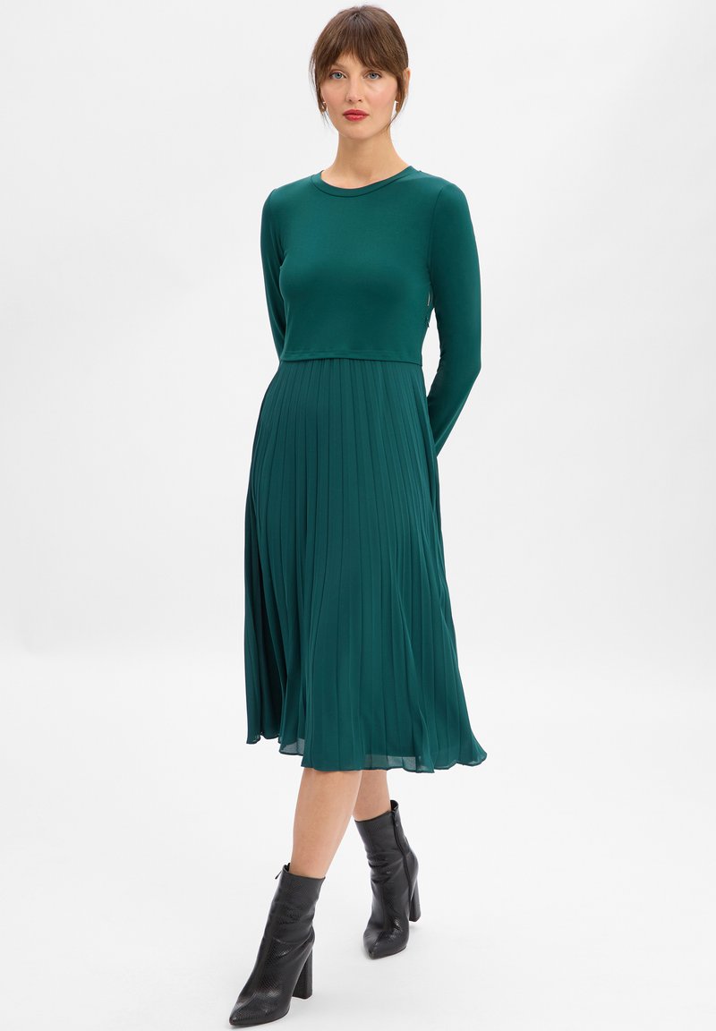 Kobieta stoi na tle białego, jednolitego tła, ubrana w długą, plisowaną sukienkę midi w kolorze morskim oraz czarne, skórzane botki na obcasie.