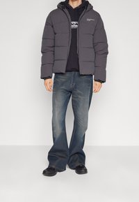 Grå pufferjakke med hætte, prydet med quiltede sektioner. Båret over en sort sweatshirt og parret med flagrende blå jeans og sorte sko.